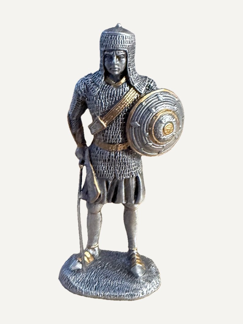 Pewter figurine of a warrior in a mail hood and mail shirt, gold-tone strap across the chest, round shield on his left arm, curved sword held point-down